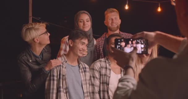 Retrato de primer plano de diversos grupos multirraciales de amigos siendo fotografiados por teléfono riendo felizmente en una fiesta divertida en una noche acogedora — Vídeos de Stock