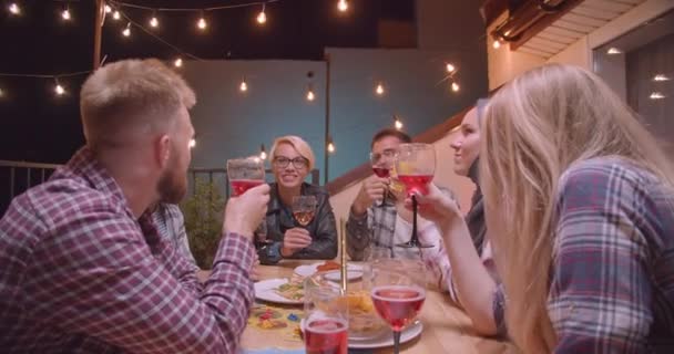 Closeup portrait of diverse multiracial group of friends drinking red wine and eating pizza relaxing in evening with fairy lights on background — Stock Video