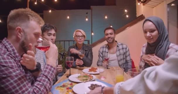 Κοντινό πορτρέτο της ποικιλόμορφης πολυφυλετικής ομάδας φίλων που πίνουν κρασί και τρώνε πίτσα χαλαρώνοντας το βράδυ με φώτα νεράιδα στο παρασκήνιο — Αρχείο Βίντεο