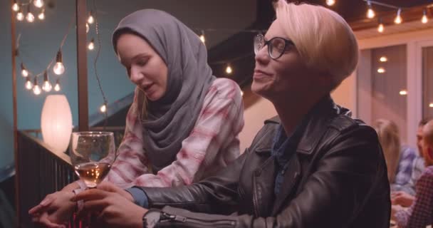 Портрет крупным планом двух женщин, разговаривающих наедине с разнообразной многорасовой группой друзей на заднем плане на вечеринке под открытым небом — стоковое видео