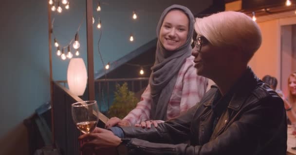 Zbliżenie portret dwóch kobiet rozmawia prywatnie z różnorodną grupą wielorasowych przyjaciół na tle na imprezie z lampami wróżek — Wideo stockowe