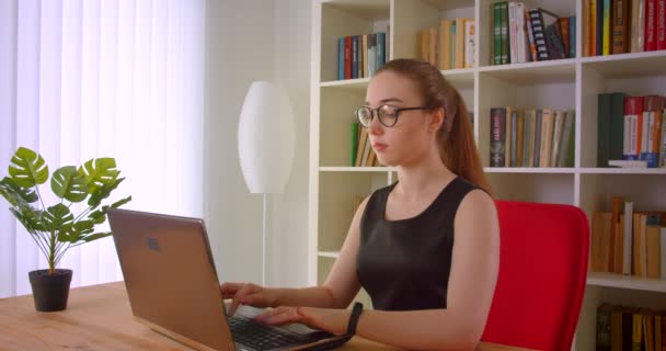 Primer plano retrato de la joven mujer de negocios pelirroja bonita en gafas usando el ordenador portátil en la oficina en el interior — Vídeo de stock