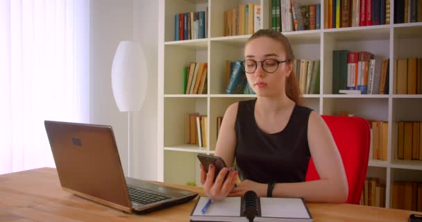 Κοντινό πλάνο της νεαρής όμορφη κοκκινομάλλα επιχειρηματίας σε γυαλιά χρησιμοποιώντας το τηλέφωνο δείχνει πράσινη οθόνη αποχρώσεων στην κάμερα κάθεται μπροστά από το laptop — Αρχείο Βίντεο