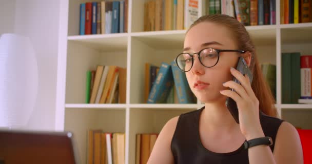 Портрет крупным планом молодой успешной рыжей предпринимательницы в очках с помощью ноутбука, имеющей телефонный звонок в офисе в помещении — стоковое видео