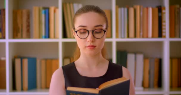 Retrato de close-up de jovem estudante ruiva bem sucedida em óculos lendo um livro na biblioteca dentro de casa com estantes em segundo plano — Vídeo de Stock