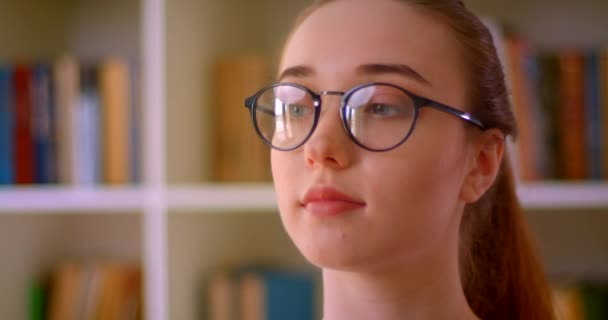 Closeup portrait of young successful redhead female student in glasses looking forward in library indoors — Stock Video