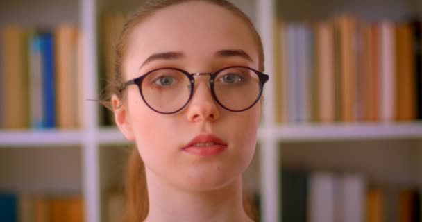 Närbild porträtt av ung vacker rödhårig kvinnlig student i glasögon tittar på kamera i biblioteket inomhus — Stockvideo