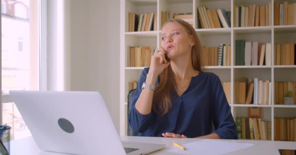 Primer plano retrato de joven atractiva mujer de negocios caucásica utilizando portátil tener conversación en el teléfono tomando notas en la oficina en el interior — Vídeos de Stock