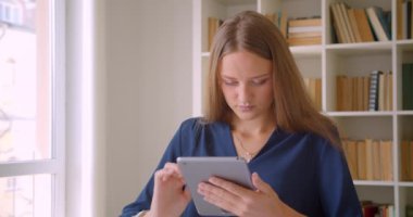 Tablet kullanan genç çekici beyaz iş kadınının kapalı ofiste masada otururken gülümseyen kameraya bakan yakın çekim portresi