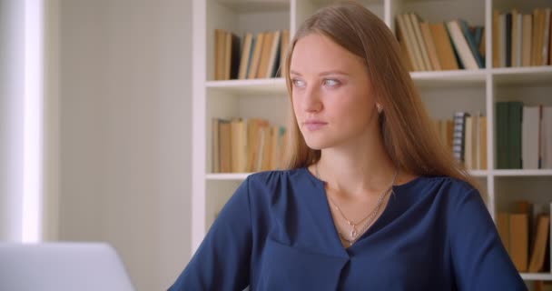 Närbild porträtt av unga kaukasiska affärskvinna tittar på kamera leende sitter vid skrivbord i kontor inomhus med bokhyllor på bakgrunden — Stockvideo