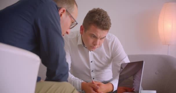 Κοντινό πορτρέτο δύο επιτυχημένων Καυκάσιων επιχειρηματιών που έχουν μια συζήτηση χρησιμοποιώντας φορητό υπολογιστή που κάθεται σε πολυθρόνες στο γραφείο σε εσωτερικούς χώρους — Αρχείο Βίντεο
