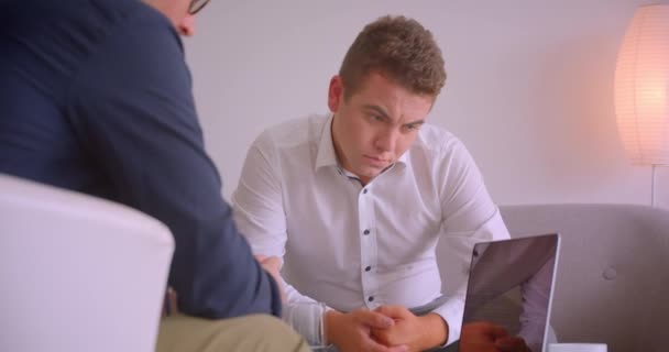 Портрет крупным планом двух успешных кавказских бизнесменов, имеющих встречу с помощью ноутбука, сидящего в креслах в офисе в помещении — стоковое видео