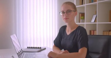 Gözlüklü genç çekici beyaz iş kadınının kapalı ofiste dizüstü bilgisayarın önünde mutlu bir şekilde gülümseyen kameraya bakan yakın çekim portresi