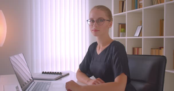 Primer plano retrato de joven atractiva mujer de negocios caucásica en gafas mirando a la cámara sentado delante de la computadora portátil en la oficina en el interior — Vídeo de stock