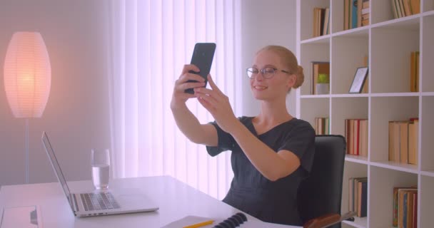 Primer plano retrato de joven atractiva mujer de negocios caucásica en gafas tomando selfies en el teléfono en la oficina en el interior — Vídeos de Stock