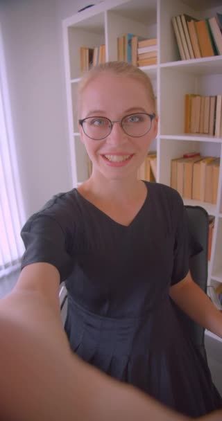 Primer plano vertical retrato de joven atractiva mujer caucásica estudiante en gafas teniendo videollamada en el teléfono sonriendo felizmente en la biblioteca en el interior — Vídeos de Stock