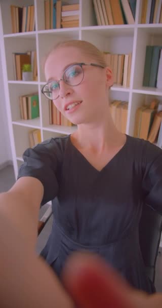 Primer plano vertical retrato de joven atractiva blogger caucásica en gafas en directo en el teléfono sonriendo alegremente en la biblioteca en el interior — Vídeos de Stock