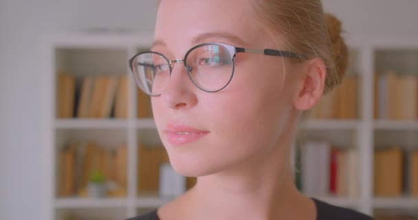 Primer plano retrato de joven atractiva estudiante caucásica en gafas girando y mirando a la cámara sonriendo felizmente en la biblioteca en el interior — Vídeos de Stock