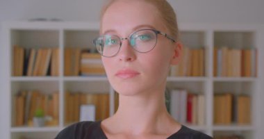 Closeup portrait of young attractive caucasian female student in glasses looking at camera in library indoors with bookshelves on background