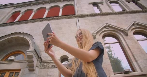 Gros plan portrait de jeune jolie femme caucasienne avec les cheveux longs dans des lunettes ayant appel vidéo sur tablette à l'extérieur dans la rue avec souffle du vent — Video