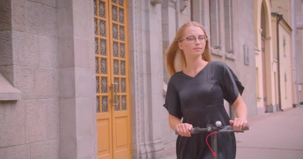 Closeup portrait of young pretty caucasian female with long hair in glasses and dress holding kick scooter looking at camera smiling outdoors — Stock Video