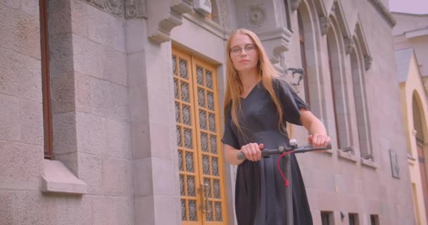 Nahaufnahme Porträt einer jungen Frau mit langen Haaren in Brille und Kleid mit Tretroller, die fröhlich in die Kamera lächelt — Stockvideo