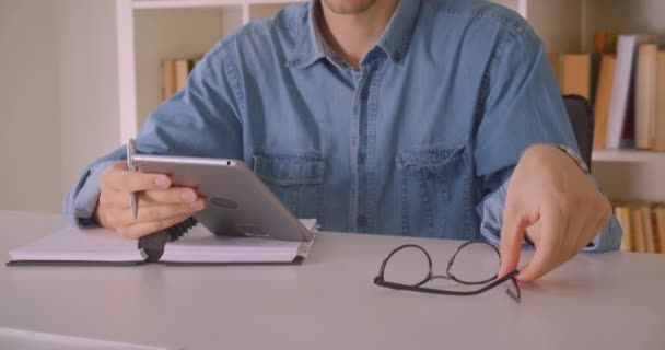Κοντινό πλάνο του νεαρού ελκυστικό Καυκάσιο αρσενικό μαθητή σε γυαλιά χρησιμοποιώντας tablet Λαμβάνοντας σημειώσεις στο γραφείο σε εσωτερικούς χώρους — Αρχείο Βίντεο