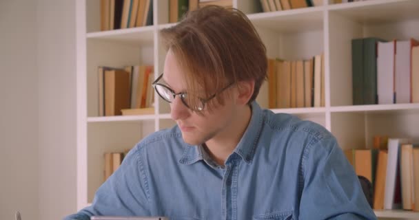 Retrato de primer plano de un atractivo estudiante caucásico en gafas usando tabletas tomando notas en la oficina en interiores — Vídeos de Stock