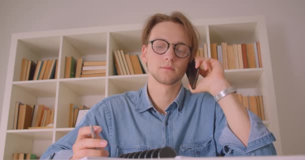 Retrato de close-up do jovem estudante masculino caucasiano atraente em óculos aprendendo a ter um telefonema tomando notas na biblioteca dentro de casa — Vídeo de Stock