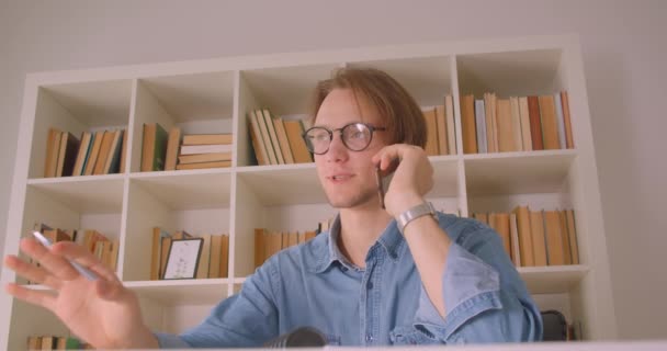 Primer plano retrato de joven guapo caucásico estudiante masculino en gafas aprendizaje tener una llamada telefónica en la biblioteca en el interior — Vídeos de Stock