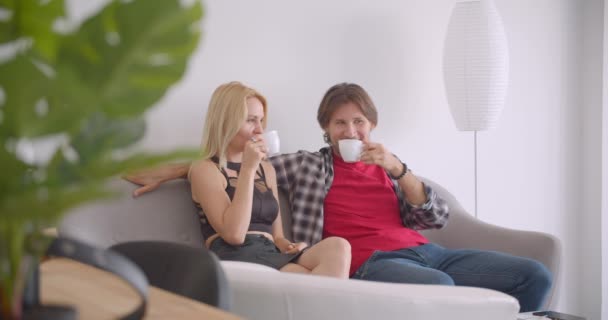 Крупним планом портрет дорослої привабливої кавказької пари п'є каву, сидячи на дивані в приміщенні в затишній квартирі — стокове відео