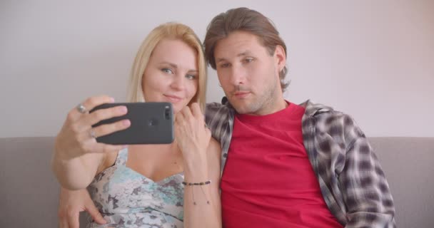 Närbild porträtt av vuxna attraktiva kaukasiska par tar selfies på telefon leende glatt sitter på soffan inomhus i lägenhet — Stockvideo