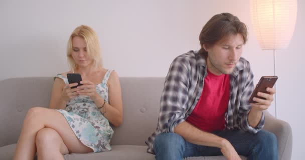 Retrato de primer plano de pareja caucásica atractiva para adultos usando teléfonos sentados en el sofá en el interior del apartamento — Vídeo de stock