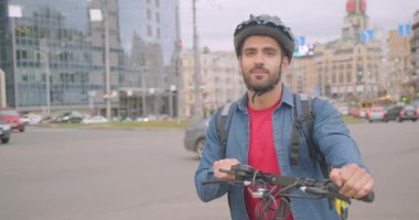 Neşeli kafkas teslimatçısının kick scooter ve sırt çantasıyla ayakta durup şehirde mutlu bir şekilde gülümseyen kameraya bakan yakın çekim portresi