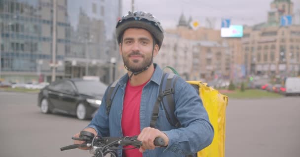 Портрет крупным планом взрослого привлекательного кавказского курьера, стоящего со скутером и рюкзаком, смотрящего в камеру в городе — стоковое видео