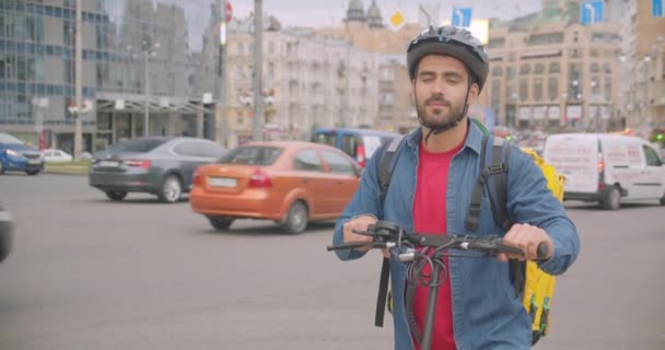 Primo piano ritratto di adulto attraente uomo di consegna caucasico in piedi con calcio scooter e zaino guardando la fotocamera sorridente felicemente in città — Video Stock