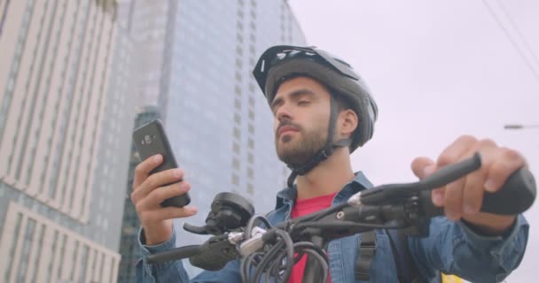 Primer plano retrato de adulto atractivo caucásico repartidor de pie con patada scooter y mochila utilizando la aplicación de navegación en el teléfono en la ciudad — Vídeos de Stock