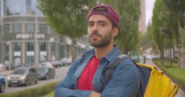 Retrato de close-up de adulto atraente homem de entrega caucasiano com mochila olhando para a câmera com os braços cruzados sobre o peito na cidade ao ar livre — Vídeo de Stock