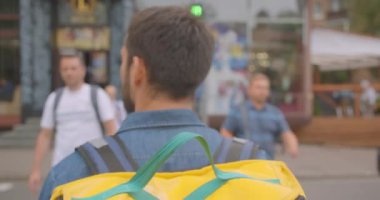 Şehir açık havada karşıdan karşıya geçen sırt çantası ile yetişkin kafkas teslimatçısının closeup geri görünümü