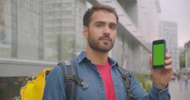 Retrato de primer plano del repartidor caucásico adulto con mochila usando el teléfono y mostrando la pantalla verde a la cámara en la calle en la ciudad al aire libre — Vídeo de stock