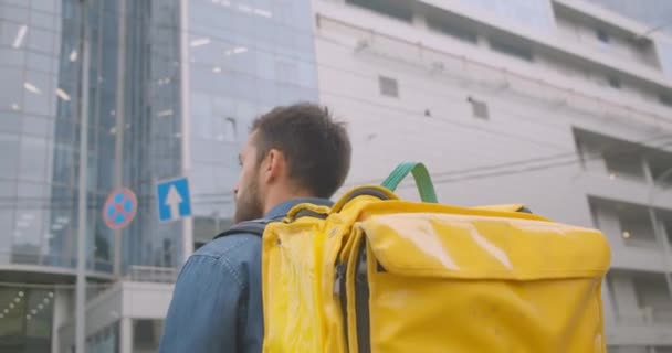 Closeup back view retrato de homem de entrega barbudo caucasiano com mochila andando na rua na cidade ao ar livre com arranha-céus — Vídeo de Stock