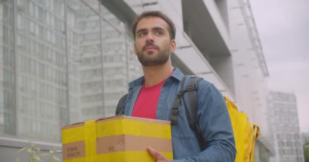 Closeup portrait of bearded caucasian delivery man with backpack holding box walking on street in city outdoors with cityscrapers — Stock Video