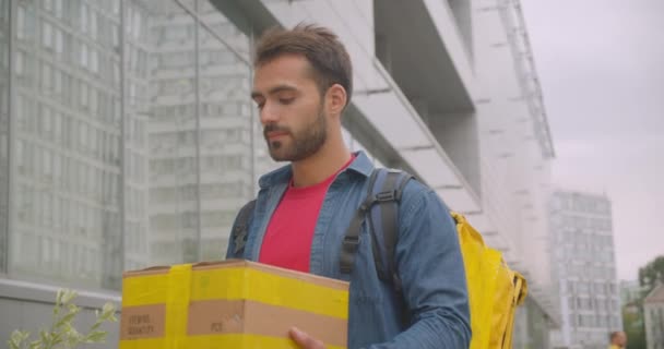 Крупним планом портрет бородатого білого чоловіка доставки з рюкзаком тримає замовлення на доставку коробки, дивлячись на камеру, посміхаючись щасливо в місті на відкритому повітрі — стокове відео