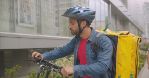 Retrato de primer plano del repartidor caucásico barbudo preparándose para enviar orden de montar patinete scooter en la ciudad al aire libre con rascacielos — Vídeo de stock