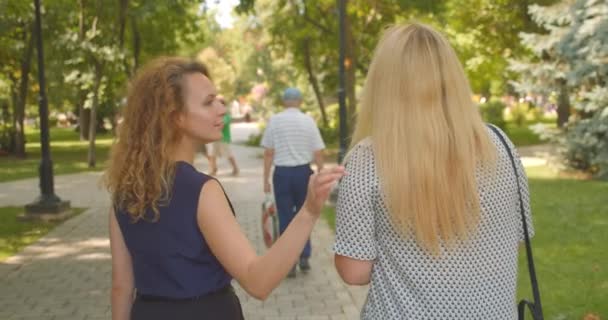 Primer plano vista trasera retrato de dos amigas caucásicas caminando en el parque al aire libre — Vídeos de Stock