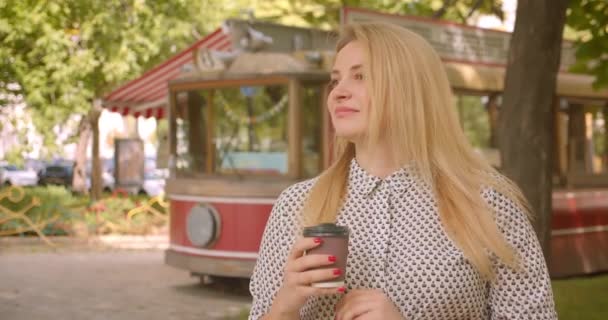 Närbild porträtt av vuxna attraktiva blonda kvinnliga hålla kaffe tittar på kamera i parken utomhus — Stockvideo