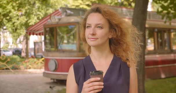 Closeup portrait of adult pretty gingerhead female looking at camera smiling cheerfully holding coffee in park outdoors — Stock Video