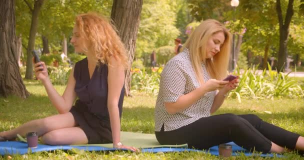 Retrato de cerca de dos amigas guapas usando teléfonos sentadas en una manta en el parque al aire libre — Vídeos de Stock