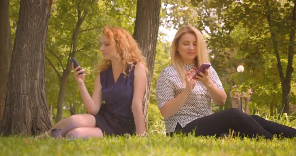 Retrato de close-up de duas amigas bonitas usando telefones compartilhando mensagens engraçadas sentadas no cobertor no parque ao ar livre — Vídeo de Stock