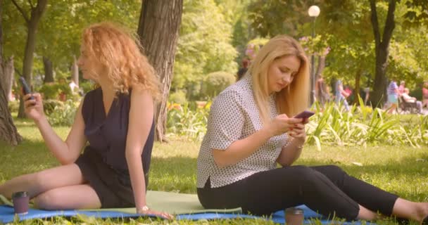 Retrato de close-up de duas amigas usando telefones sentados em cobertor no parque ao ar livre — Vídeo de Stock